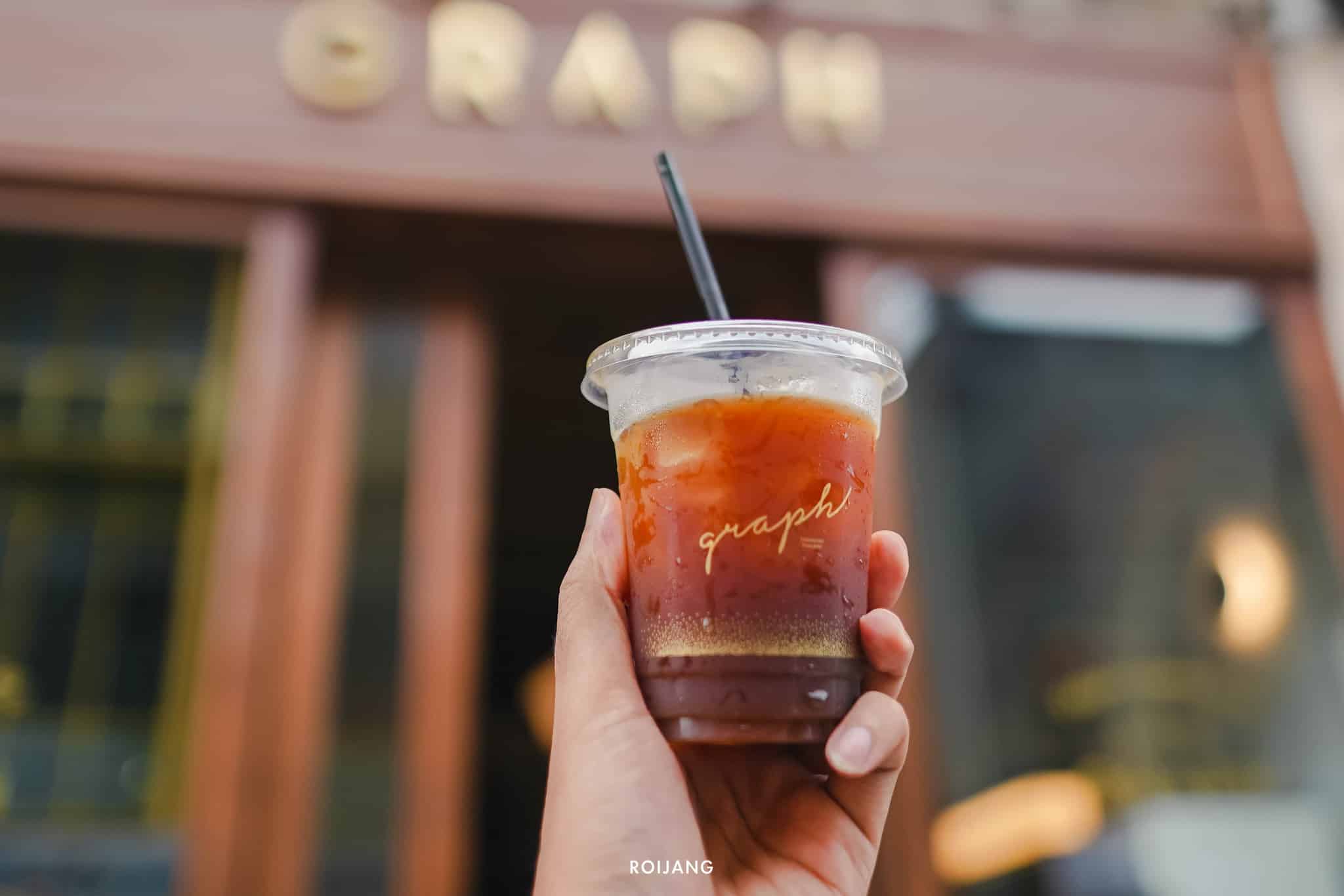มือหนึ่งถือแก้วพลาสติกใส่น้ำแข็งกาแฟพร้อมหลอดอยู่ด้านหน้า คาเฟ่ภูเก็ต โดยมีป้ายชื่อ "GRAPH" อยู่เหนือทางเข้า.