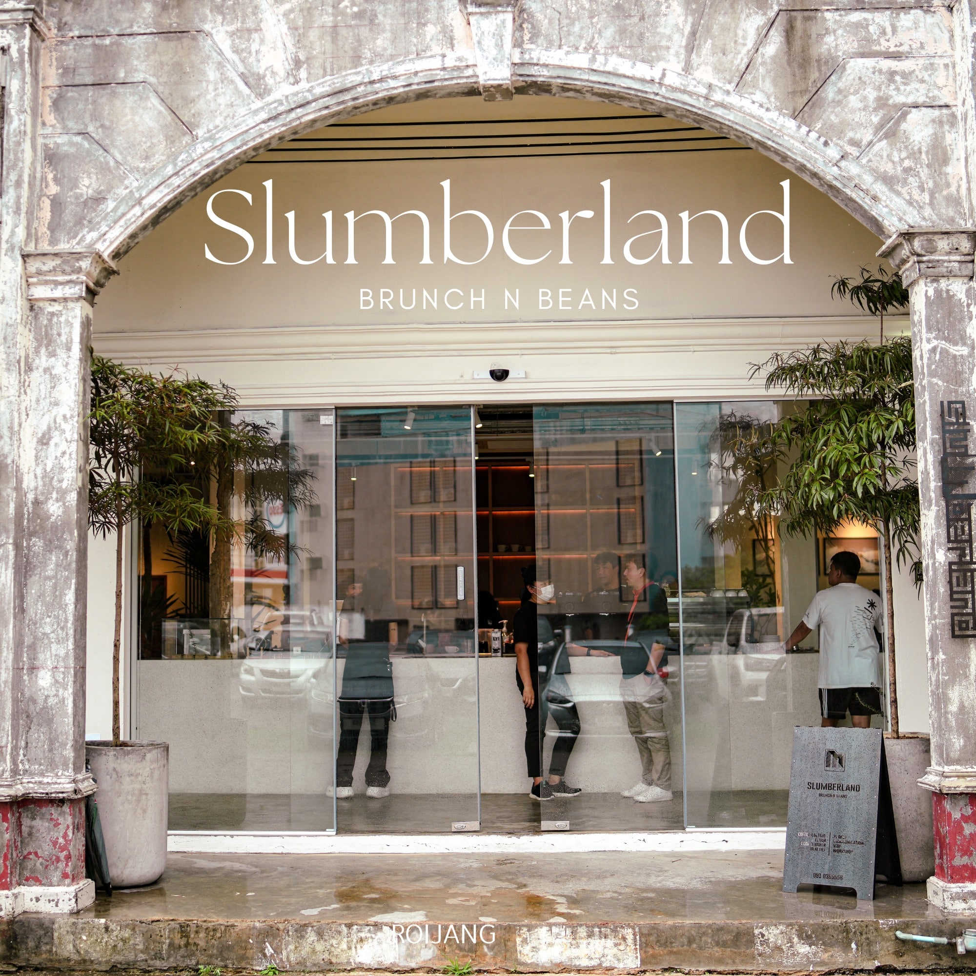 ทางเข้าของ Slumberland Brunch N Beans คาเฟ่ภูเก็ต มีประตูกระจก คนภายใน และต้นไม้ในกระถางอยู่ทั้งสองข้างของทางเข้า.