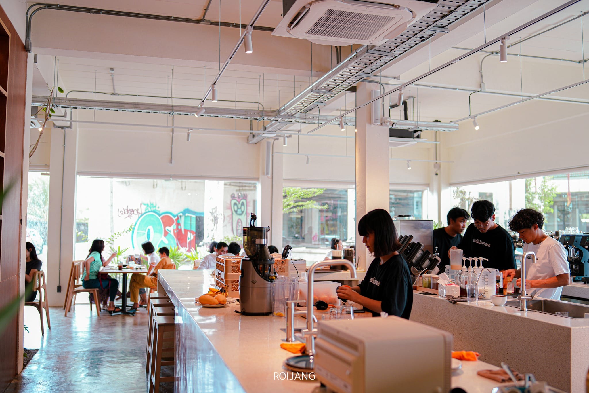 หลายคนทำงานอยู่หลังเคาน์เตอร์เตรียมเครื่องดื่มใน คาเฟ่ภูเก็ต ที่ทันสมัยและสว่างไสว ขณะที่ลูกค้าได้นั่งอยู่ที่โต๊ะใกล้หน้าต่างขนาดใหญ่ในฉากหลัง.