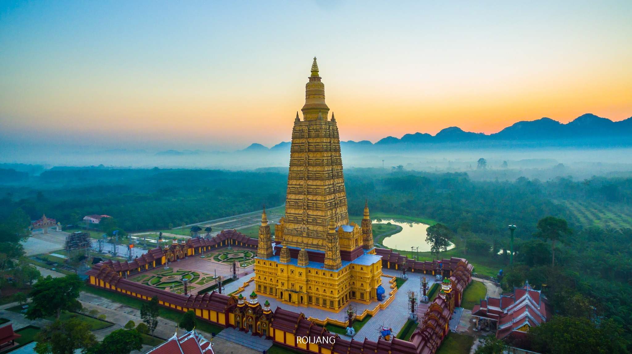 ภาพมุมสูงของวัดสีทองขนาดใหญ่ที่ล้อมรอบด้วยสวนและกำแพงในยามพระอาทิตย์ขึ้น โดยมีภูเขาและหมอกเป็นฉากหลัง—สถานที่ท่องเที่ยวที่น่าหลงใหลในกระบี่สำหรับผู้ที่ต้องการค้นพบความงามของกระบี่.