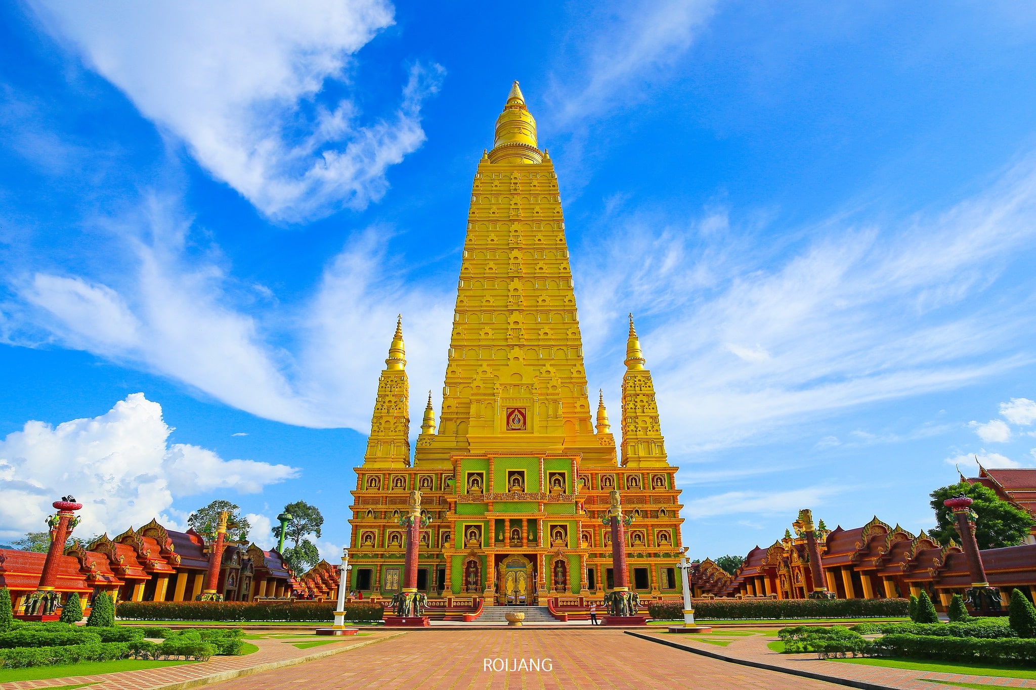 เจดีย์พุทธสีทองขนาดใหญ่ที่มีรายละเอียดประณีตตั้งอยู่ใต้ท้องฟ้าสีคราม ขนาบข้างด้วยอาคารที่สมมาตรและสวนสีเขียวชอุ่ม—สถานที่ที่ไม่ควรพลาดสำหรับผู้ที่มาเที่ยวกระบี่หรือกำลังมองหาสถานที่ท่องเที่ยวในกระบี่.