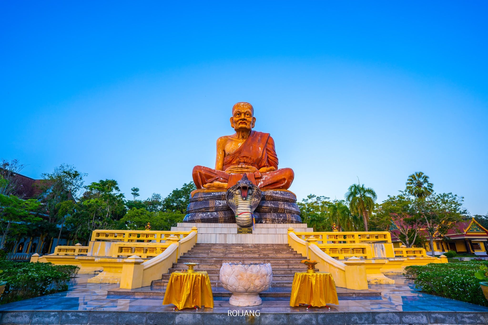 รูปปั้นสัมฤทธิ์ขนาดใหญ่ของพระสงฆ์นั่งบนแท่นยกสูง ล้อมรอบด้วยต้นไม้และรั้วสีเหลืองภายใต้ท้องฟ้าสีฟ้าใส—สถานที่เงียบสงบสำหรับผู้ที่ต้องการเที่ยวกระบี่และค้นพบสถานที่ท่องเที่ยวที่ไม่เหมือนใครในกระบี่.