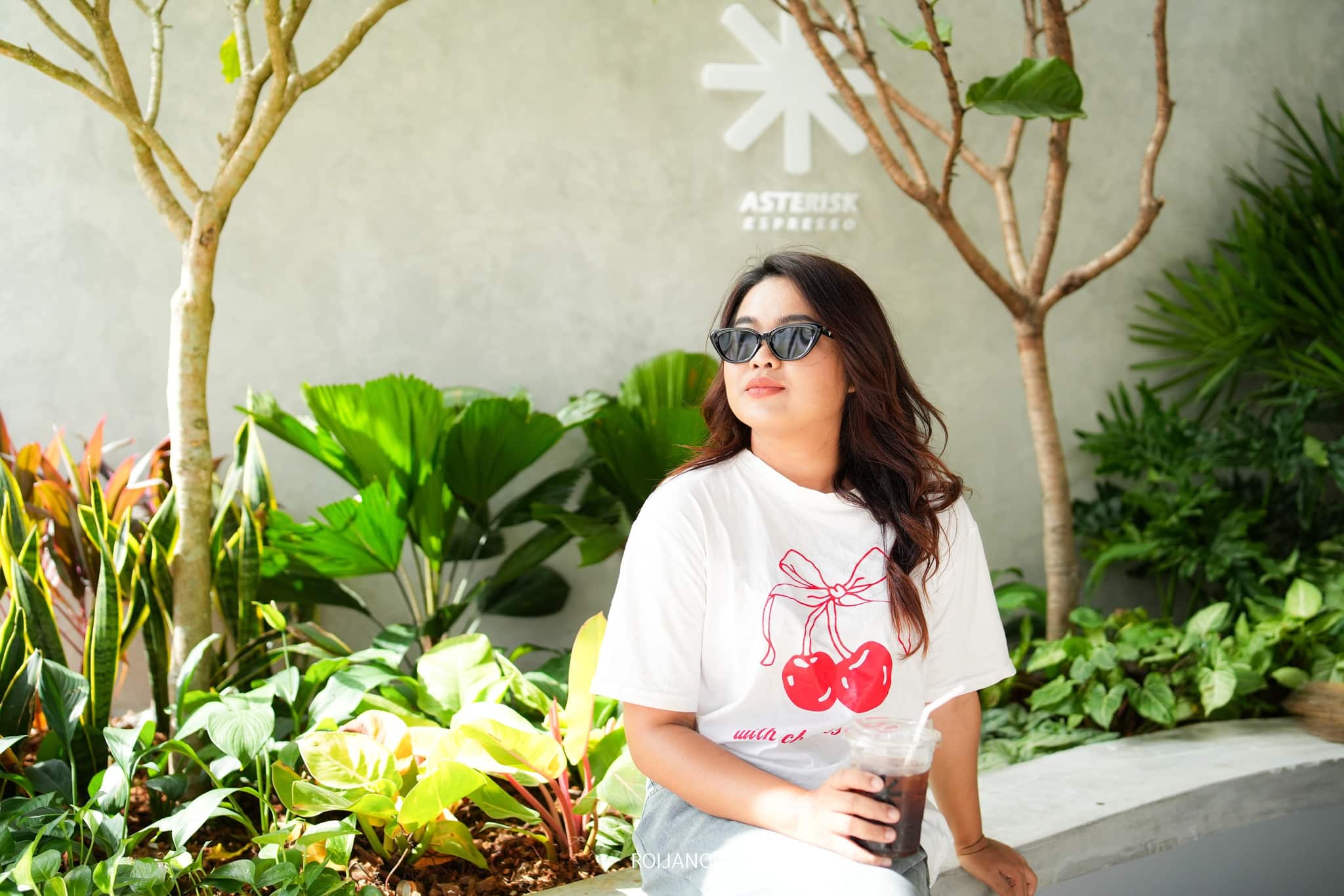 ผู้หญิงคนหนึ่งสวมแว่นกันแดดและเสื้อยืดสีขาวลายเชอร์รี่ นั่งอยู่บนม้านั่งคอนกรีต ถือแก้วพลาสติก รวม ร้านกาแฟภูเก็ต หลากหลายสไตล์ คาเฟ่ภูเก็ต ดังสุดชิคในเกาะสวรรค์ ดื่มด่ำกับกลิ่นกาแฟหอมๆ และบรรยากาศ จิบกาแฟชมวิวพระอาทิตย์ คาเฟ่ที่ไม่ลับอีกต่อไป ท่ามกลางพืชพรรณเขียวชอุ่ม ที่ร้านกาแฟ Asterisk Espresso ในภูเก็ต ซึ่งเป็นคาเฟ่ที่มีชื่อเสียงในเรื่องบรรยากาศที่ผ่อนคลาย.