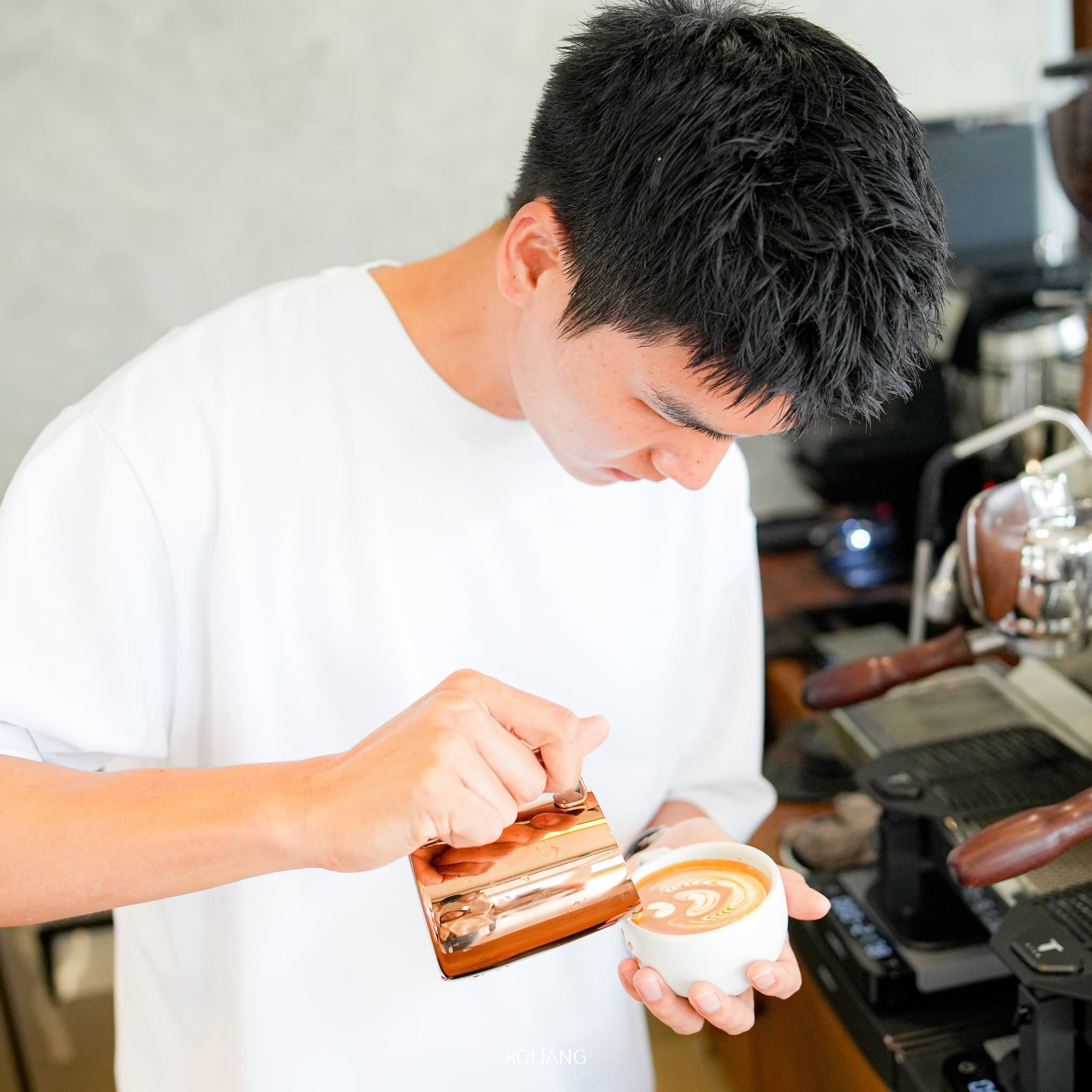 ชายในเสื้อเชิ้ตสีขาวค่อยๆ เทนมร้อนลงในถ้วยกาแฟอย่างระมัดระวัง สร้างลวดลายลาเต้อาร์ตข้างเครื่องเอสเพรสโซใน ร้านกาแฟภูเก็ต ที่แสนอบอุ่น.