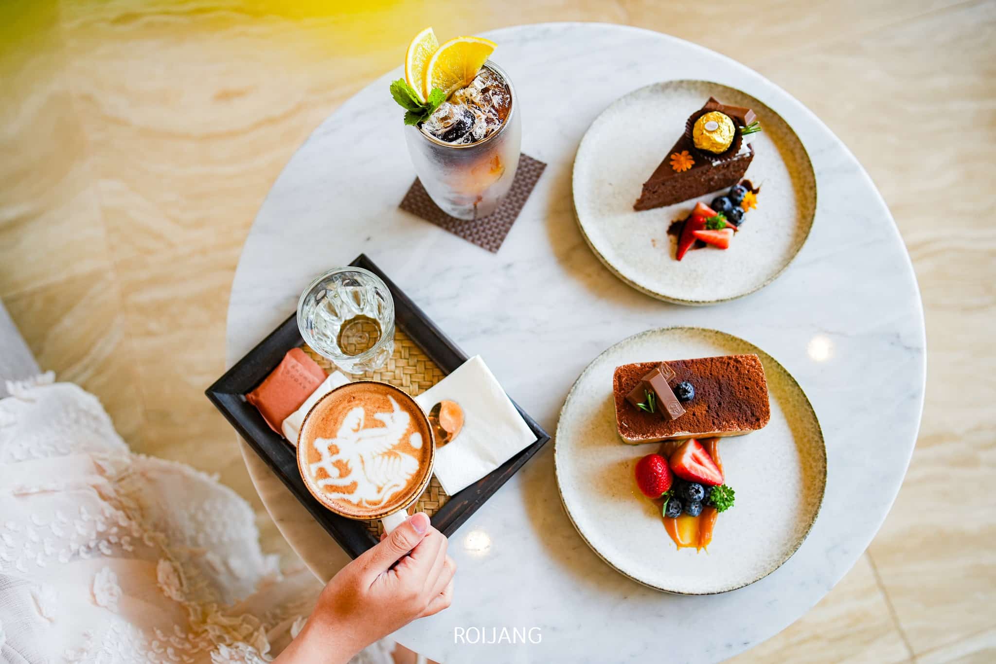บุคคลหนึ่งนั่งอยู่ที่โต๊ะหินอ่อนใน ร้านกาแฟภูเก็ต พร้อมถาดที่มีลาเต้อาร์ต แก้วน้ำเปล่า คุกกี้ จานเค้กช็อกโกแลตที่โรยด้วยเบอร์รี่ และเครื่องดื่มเย็นสดชื่นที่ตกแต่งด้วยส้มฝานหนึ่งชิ้น.