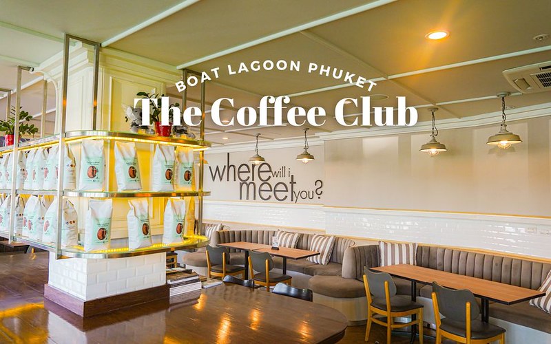 ภายในร้าน The Coffee Club ที่ Boat Lagoon, คาเฟ่ภูเก็ต โดดเด่นด้วยที่นั่งทันสมัย โคมไฟแขวน และถุงกาแฟบนชั้นวาง ข้อความบนภาพประกอบด้วยชื่อร้านและ "เราจะพบคุณที่ไหน?" — คาเฟ่ภูเก็ตที่ต้องไปเยือน สำหรับคนรักกาแฟ.