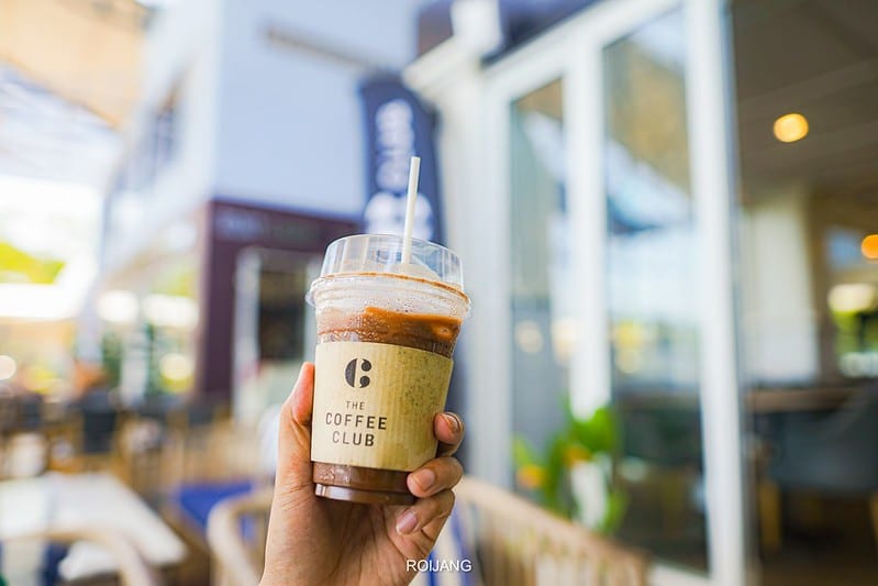 มือหนึ่งถือแก้วพลาสติกใส่น้ำแข็งกาแฟพร้อมหลอด มีป้ายเขียนว่า "The Coffee Club" อยู่ด้านหน้าฉาก คาเฟ่ภูเก็ต ที่เบลอ.