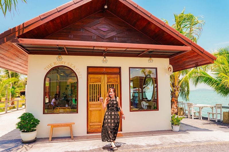 ผู้หญิงในชุดสีดำยืนอยู่หน้าร้าน The Ruby Sea Café คาเฟ่ภูเก็ต ที่มีเสน่ห์ พร้อมต้นปาล์มและที่นั่งกลางแจ้งริมน้ำ.