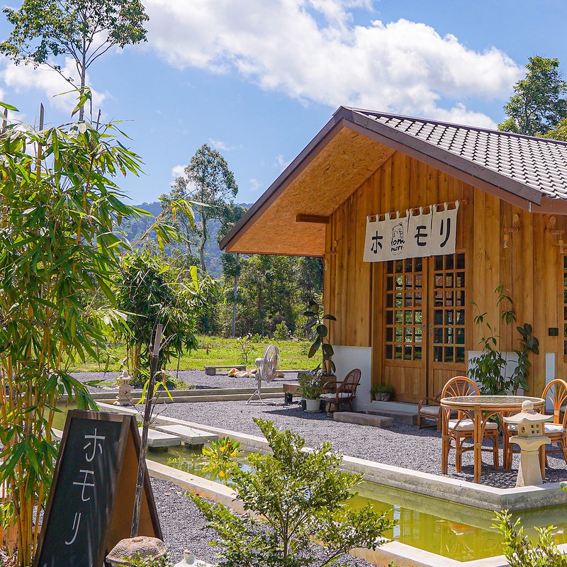 คาเฟ่ภูเก็ต ไม้ขนาดเล็กที่มีหลังคากระเบื้องตั้งอยู่ข้างสระน้ำและที่นั่งกลางแจ้ง ล้อมรอบด้วยต้นไม้สีเขียวภายใต้ท้องฟ้าสีคราม มีป้ายที่มีตัวอักษรภาษาญี่ปุ่นตั้งอยู่ใกล้ทางเข้า.