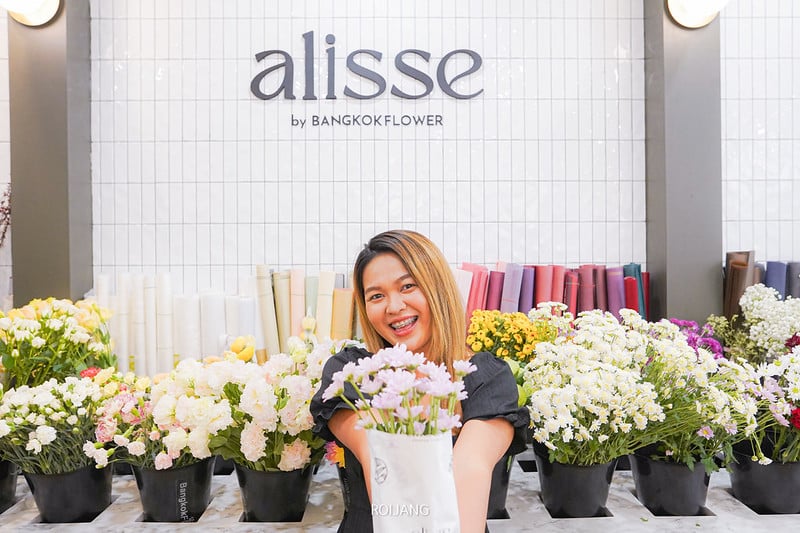 ผู้หญิงคนหนึ่งยิ้มขณะถือช่อดอกไม้ไว้ข้างหน้าการจัดแสดงดอกไม้ที่ Alisse by Bangkok Flower สถานที่น่ารักใกล้ คาเฟ่ภูเก็ต.