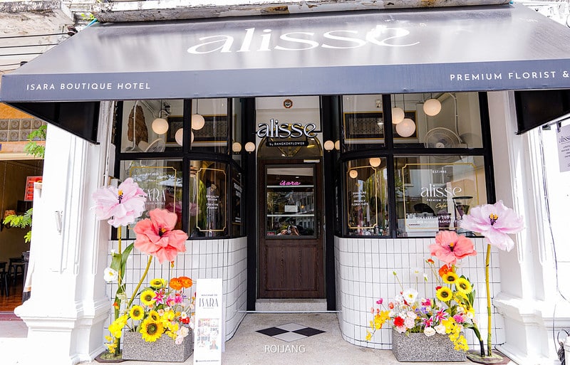 หน้าร้านของ "alissé" มีกันสาดสีดำ ประตูกระจก ผนังด้านหน้าปูกระเบื้องสีขาว และจัดดอกไม้ขนาดใหญ่ไว้สองข้างทางเข้า— ร้านกาแฟภูเก็ต ที่ชวนให้หลงใหล เหมาะสำหรับคนรักกาแฟ.