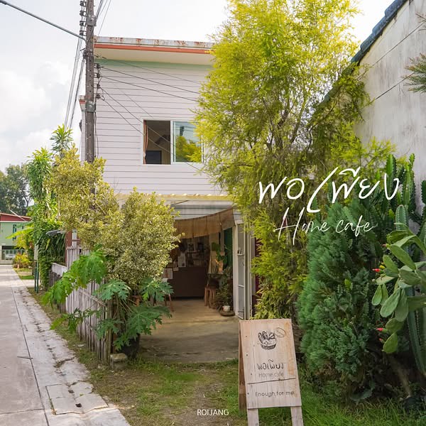 คาเฟ่ภูเก็ต ขนาดเล็กที่มีป้ายไม้ตั้งอยู่หน้าทางเข้า ล้อมรอบด้วยต้นไม้เขียวขจีบนถนนแคบ โดยมีอาคารสองชั้นตั้งอยู่ด้านหลัง.