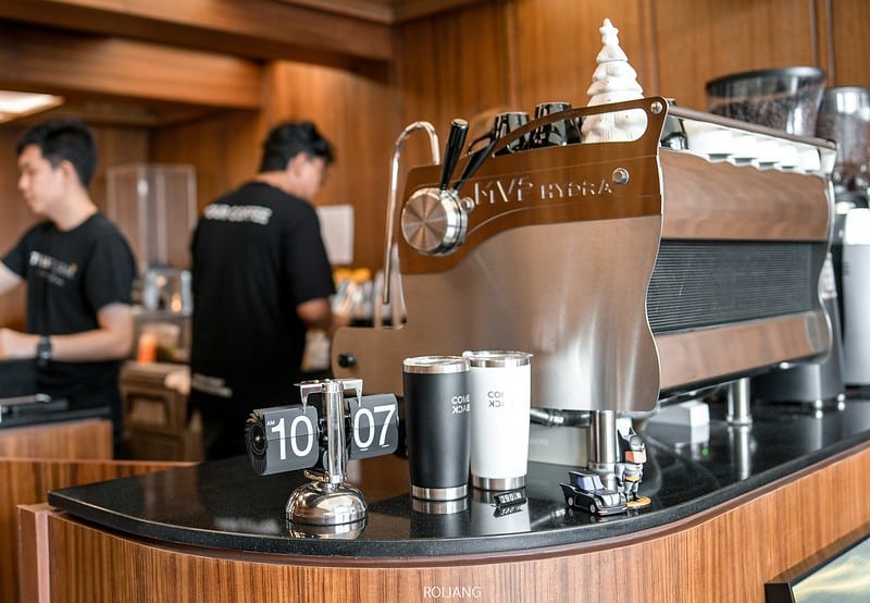 คาเฟ่ภูเก็ต สมัยใหม่ พร้อมเครื่องชงกาแฟเอสเพรสโซขนาดใหญ่ แก้วเดินทางสองใบ นาฬิกาตั้งโต๊ะแบบพลิกแสดงเวลา 10:07 และบาริสต้าสองคนกำลังทำงานอยู่เบื้องหลัง.