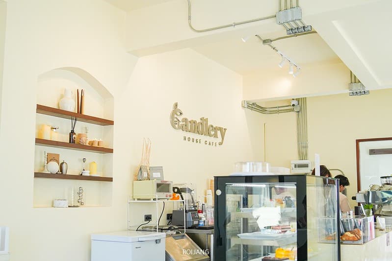 ภายใน คาเฟ่ภูเก็ต ที่สว่างไสวและเรียบง่าย มีชั้นวางของตกแต่ง, ตู้โชว์ขนมหวาน, และป้ายที่เขียนว่า "Canderly House Cafe" บนผนังสีครีม.