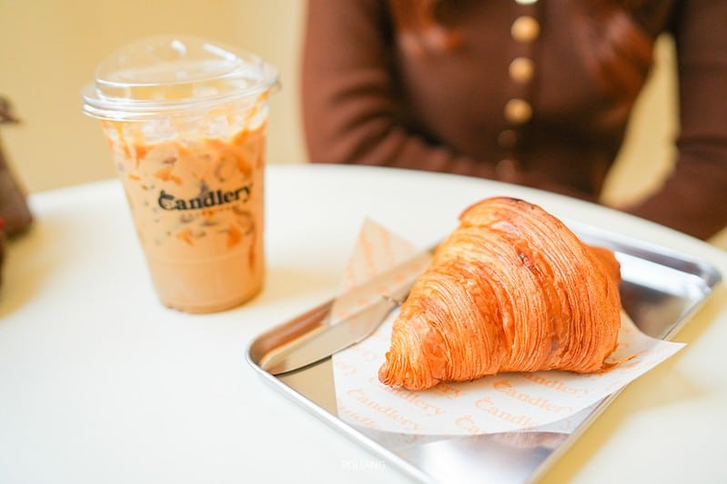 ครัวซองต์ขนาดใหญ่บนถาดโลหะและกาแฟเย็นในแก้วพลาสติกวางอยู่บนโต๊ะสีขาวใน คาเฟ่ภูเก็ต ; มีคนนั่งอยู่ด้านหลัง.