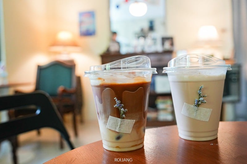 แก้วพลาสติกสองใบที่บรรจุเครื่องดื่มกาแฟเย็นพร้อมฝาปิดวางอยู่บนโต๊ะไม้ภายใน คาเฟ่ภูเก็ต ที่อบอุ่นและแสงไฟนวลตา.