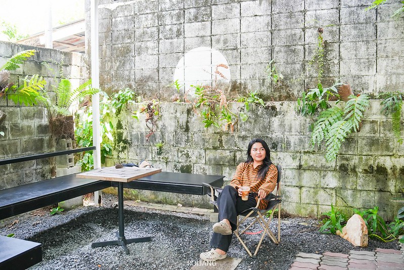 ผู้หญิงคนหนึ่งนั่งอยู่บนเก้าอี้พับในลานกลางแจ้งของ ร้านกาแฟภูเก็ต ล้อมรอบด้วยกำแพงหิน พืชสีเขียว โต๊ะ และพื้นกรวด พร้อมถือถ้วยในมือ.