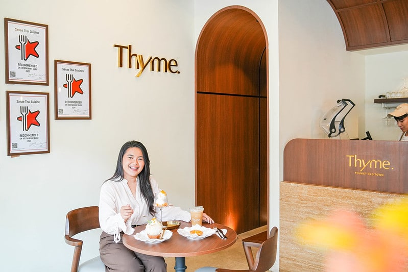 ผู้หญิงคนหนึ่งนั่งอยู่ที่โต๊ะพร้อมของหวานและเครื่องดื่มภายใน Thyme คาเฟ่ภูเก็ต โดยมีรางวัลแสดงอยู่บนผนังและพนักงานคนหนึ่งอยู่ที่เคาน์เตอร์ในพื้นหลัง.