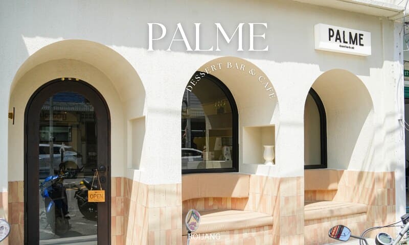 ภายนอกของ PALME dessert bar and café ( คาเฟ่ภูเก็ต ) พร้อมหน้าต่างโค้ง ประตูที่มีป้าย "เปิด" และผนังสีขาวที่มีชื่อคาเฟ่.
