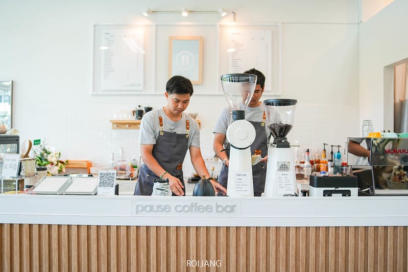 บาริสต้าสองคนในผ้ากันเปื้อนสีเทาเตรียมเครื่องดื่มอยู่หลังเคาน์เตอร์ที่ Pause Coffee Bar ร้านกาแฟภูเก็ต ที่สว่างไสวและทันสมัย พร้อมอุปกรณ์กาแฟและเมนูที่มองเห็นได้ในพื้นหลัง.