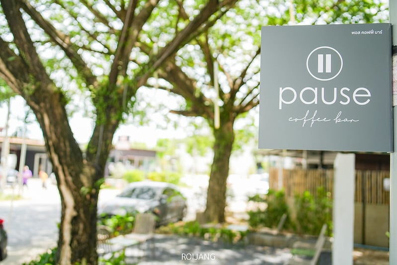 ป้ายสี่เหลี่ยมผืนผ้าสำหรับ "Pause coffee bar" แขวนอยู่ภายนอกใกล้ต้นไม้ใบเขียวและถนนที่มีแสงแดดส่องเชิญชวนผู้มาเยือนสู่ คาเฟ่ภูเก็ต ที่มีเสน่ห์แห่งนี้.