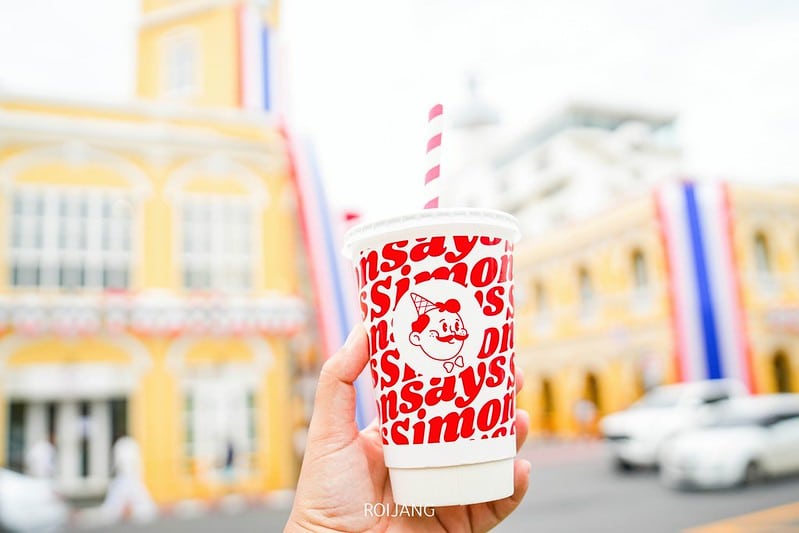 มือหนึ่งถือแก้วเครื่องดื่มที่มีข้อความสีแดงเขียนว่า "Simon" อยู่ด้านหน้าอาคารสีเหลืองซึ่งมีหน้าต่างโค้งและประดับตกแต่งด้วยสีแดง ขาว และน้ำเงิน สะท้อนเสน่ห์อันมีชีวิตชีวาของ คาเฟ่ภูเก็ต.