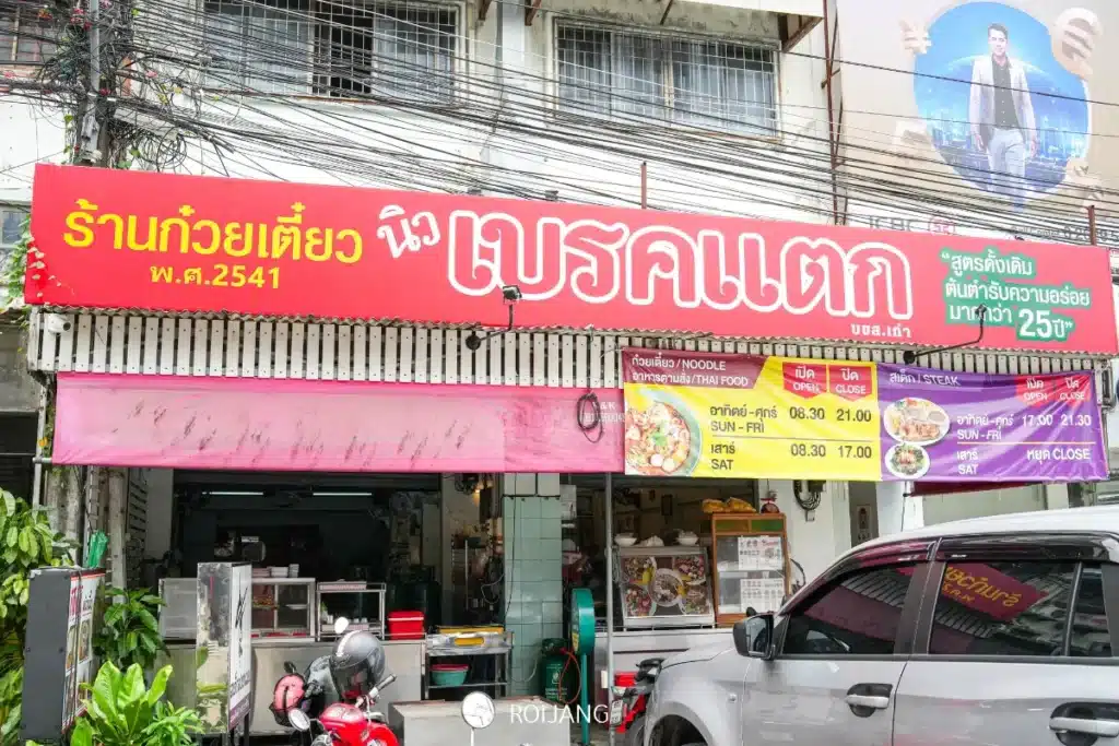 ภาพถนนหน้าร้านก๋วยเตี๋ยวไทยที่มีป้ายสีแดงติดอยู่เหนือทางเข้า แสดงรายการอาหารและราคาเป็นภาษาไทย มีรถจักรยานยนต์จอดอยู่ด้านหน้า และมีส่วนขายขนมอบรสเลิศอยู่ภายในร้าน.