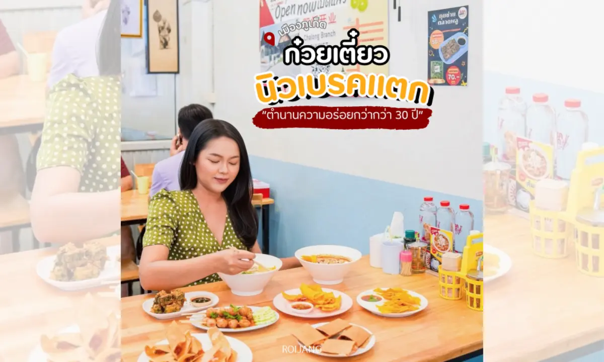 ผู้หญิงคนหนึ่งนั่งอยู่ที่โต๊ะในร้านอาหาร กำลังกินก๋วยเตี๋ยว โดยมีจานอาหารหลายจานล้อมรอบ—รวมถึงของอบพิเศษที่ดูหรูหรา—พร้อมด้วยเมนูอาหารสีสันสดใสบนผนังและเครื่องปรุงรสต่างๆ เป็นฉากหลัง.
