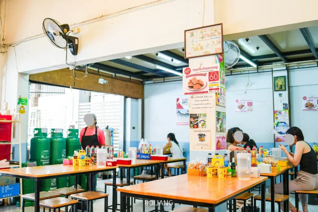 ภายในร้านอาหารที่สว่างและสบายๆ มีโต๊ะไม้ เครื่องปรุงรส และลูกค้าที่นั่งอยู่ โปสเตอร์และเมนู—บางอันเน้นสินค้าเบเกอรี่ชั้นดี—ติดอยู่บนผนังและเสา.