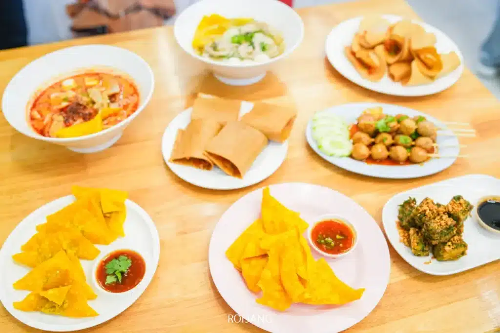 โต๊ะไม้ที่มีอาหารไทยหลากหลายชนิด รวมถึงก๋วยเตี๋ยวเรือแตก, ซุป, วอนตันทอด, ปอเปี๊ยะทอด, ย่างเสียบไม้, และน้ำจิ้ม, ทั้งหมดจัดเรียงอย่างสวยงามบนจานขาว.