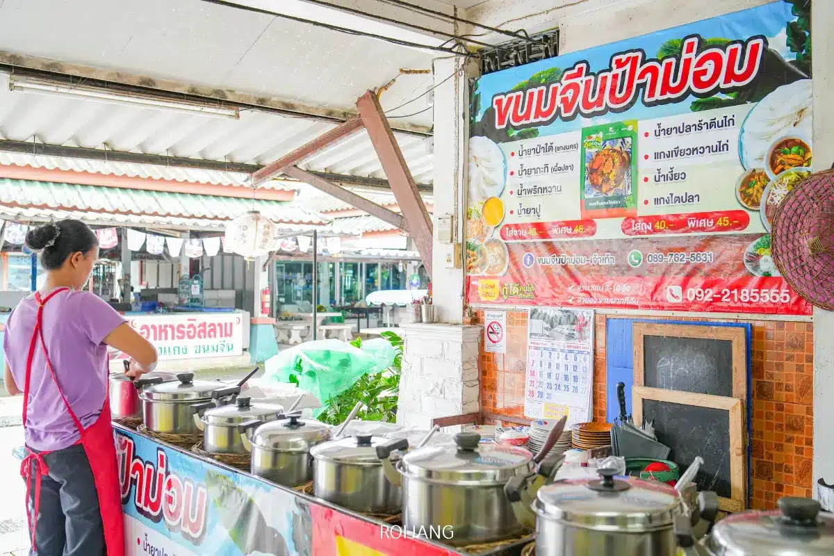 ผู้หญิงคนหนึ่งสวมผ้ากันเปื้อนยืนอยู่หลังแถวหม้ออาหารที่ร้านขายอาหารไทย ขนมจีนป้าม่อม กลางแจ้ง โดยมีเมนูอาหารหลากสีสันติดแสดงอยู่บนผนัง.
