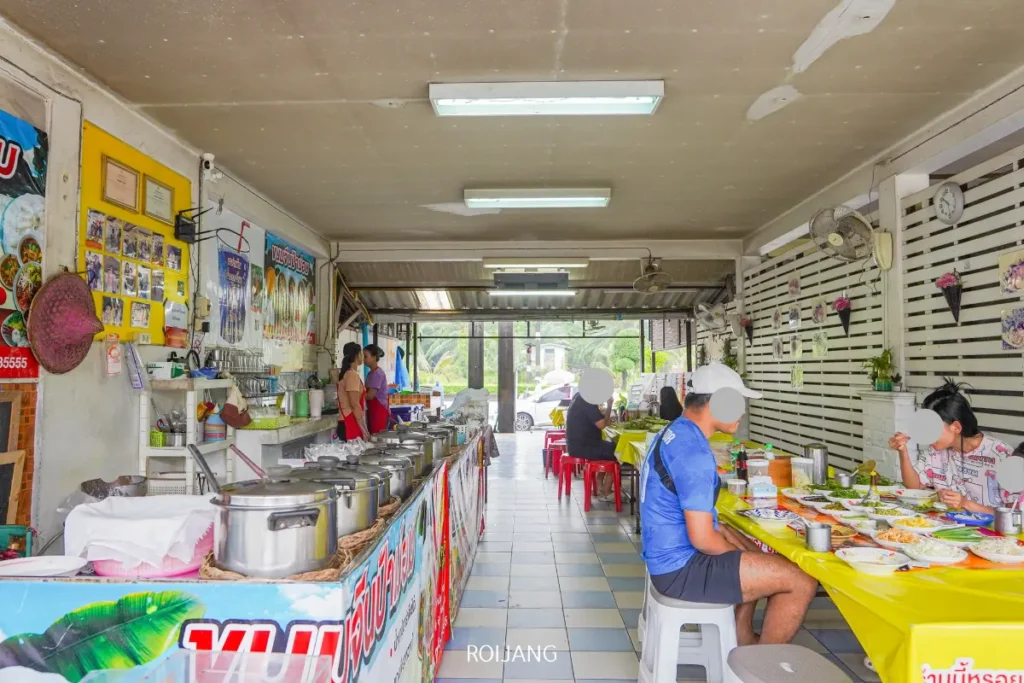 ชายคนหนึ่งนั่งอยู่ที่โต๊ะในร้านอาหาร พร้อมที่จะเพลิดเพลินกับ ขนมจีนป้าม่อม ที่มีชื่อเสียง.