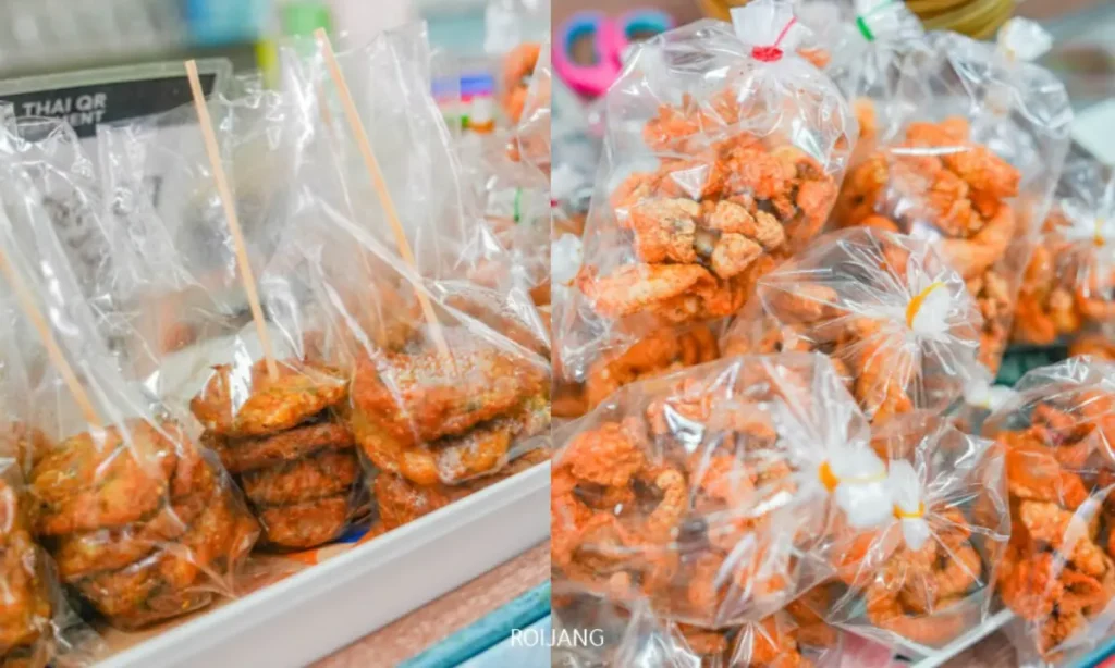 ถุงพลาสติกใสที่บรรจุชิ้นส่วนของอาหารทอดปรุงรสถูกจัดแสดงบนแผงขายของในตลาดข้าง ขนมจีนป้าม่อม บางถุงปิดผนึกไว้ บางถุงมีไม้เสียบ.