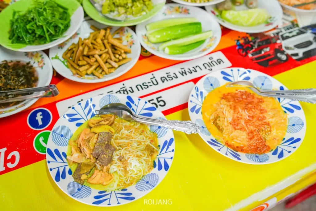 สองจานของก๋วยเตี๋ยวแกงไทยที่รู้จักกันในชื่อ ขนมจีนป้าม่อม พร้อมช้อนวางอยู่บนโต๊ะสีแดงและเหลือง ล้อมรอบด้วยเครื่องเคียงผักและผักดอง.