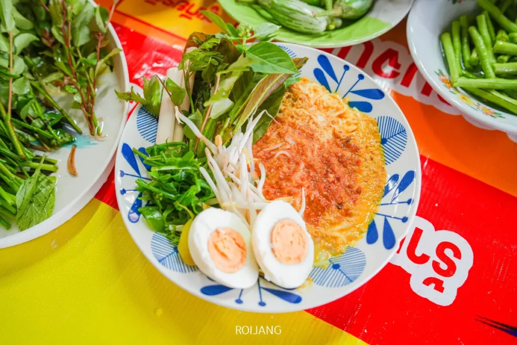 จานที่มีไข่เจียวสุก, ไข่ต้มผ่าครึ่ง, สมุนไพรสด, และถั่วงอกวางอยู่บนโต๊ะสีสันสดใสข้างๆ ขนมจีนป้าม่อม และเครื่องเคียงผักต่างๆ.