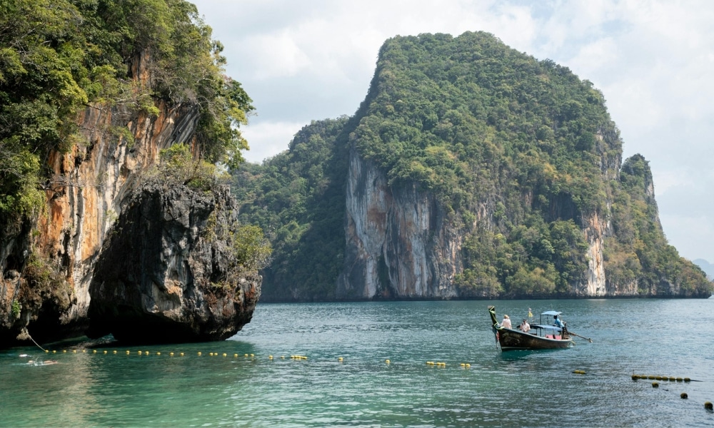 เรือไม้ลำหนึ่งลอยอยู่บนผืนน้ำสีฟ้าใสราวเทอร์ควอยซ์ ใกล้หน้าผาหินและป่าไม้ที่ปกคลุมไปด้วยต้นไม้ใหญ่ รวมถึงเกาะหินปูนขนาดมหึมา ท่ามกลางท้องฟ้าที่มีเมฆลอยบางเป็นบางช่วง—เหมาะอย่างยิ่งสำหรับผู้ที่กำลังมองหากิจกรรมท่องเที่ยวในกระบี่.