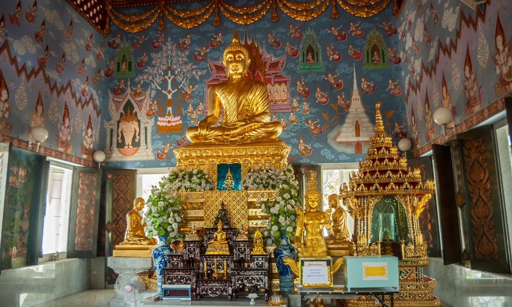 พระพุทธรูปทองคำบนแท่นบูชาที่ประดับประดาอย่างวิจิตร พร้อมด้วยเครื่องบูชาและของตกแต่งต่างๆ ล้อมรอบด้วยผนังภาพจิตรกรรมฝาผนังสีสันสดใสและลวดลายที่ซับซ้อนภายในวัด—เป็นจุดเด่นที่สร้างแรงบันดาลใจสำหรับทุกคนที่วางแผนจะ เที่ยวกระบี่.