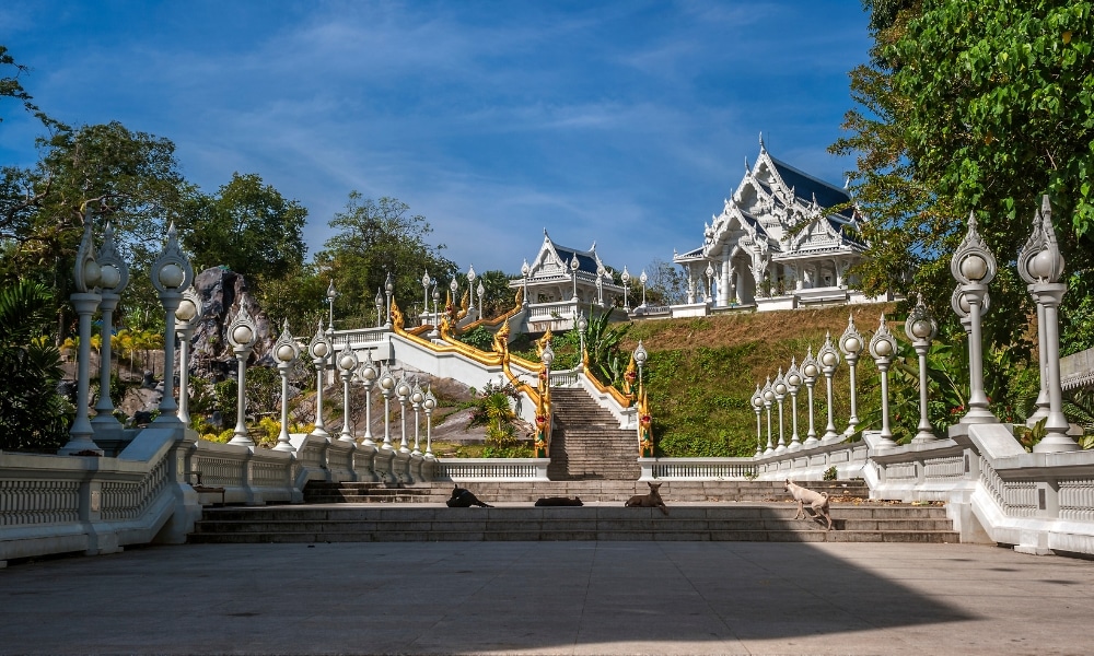บันไดกว้างที่มีราวจับประดับอย่างวิจิตรและรูปปั้นทองคำนำขึ้นไปยังวัดสีขาวที่มีหลังคาแหลม—หนึ่งในสถานที่ท่องเที่ยวที่ต้องไปเยือนในกระบี่—ล้อมรอบด้วยต้นไม้ภายใต้ท้องฟ้าสีคราม.