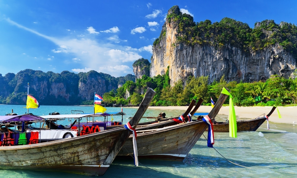 เรือไม้แบบดั้งเดิมที่มีธงสีสันสดใสจอดทอดสมออยู่ในน้ำตื้นบนชายหาดเขตร้อน—เป็นภาพอันเป็นเอกลักษณ์สำหรับผู้ที่ต้องการ เที่ยวกระบี่ พร้อมด้วยหน้าผาหินปูนและพืชพรรณเขียวขจีที่โอบล้อมทิวทัศน์อันมีชีวิตชีวาภายใต้ท้องฟ้าสีคราม.