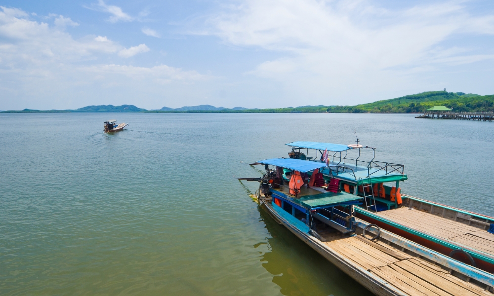 เรือไม้สองลำลอยอยู่บนน้ำสงบใกล้ชายฝั่ง ท่ามกลางทิวทัศน์เขียวขจีและภูเขาในเบื้องหลัง—ภาพที่ชวนให้ใครก็ตามที่อยากเที่ยวกระบี่ภายใต้ท้องฟ้าที่มีเมฆบางส่วนมาเยือน.