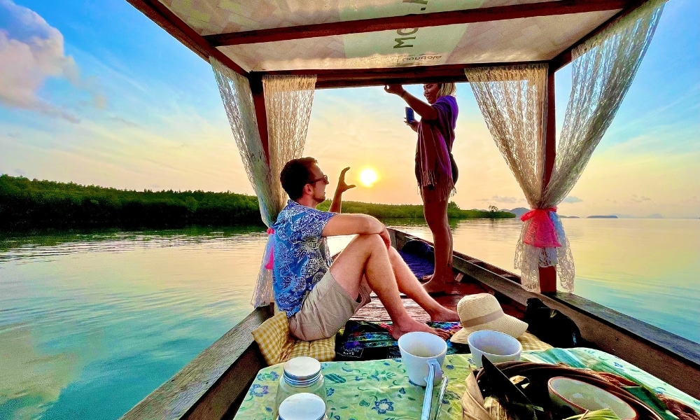 สองคนบนเรือไม้ในยามพระอาทิตย์ตกดินที่ เที่ยวกระบี่—คนหนึ่งนั่งในขณะที่อีกคนยืนถือเครื่องดื่ม ถ้วยและหมวกวางอยู่บนโต๊ะในฉากหน้า ขณะที่น้ำสงบล้อมรอบทัศนียภาพอันเงียบสงบของ ที่เที่ยวกระบี่.