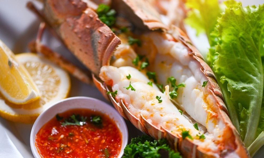 หางกุ้งมังกรเสิร์ฟในเปลือกพร้อมสมุนไพรสับ เสิร์ฟพร้อมกับมะนาวฝาน ผักกาดหอม และถ้วยเล็กซอสจิ้มสีแดง—เมนูที่ต้องลองเมื่อมาเที่ยวกระบี่เพื่อลิ้มรสอาหารทะเลสดใหม่.