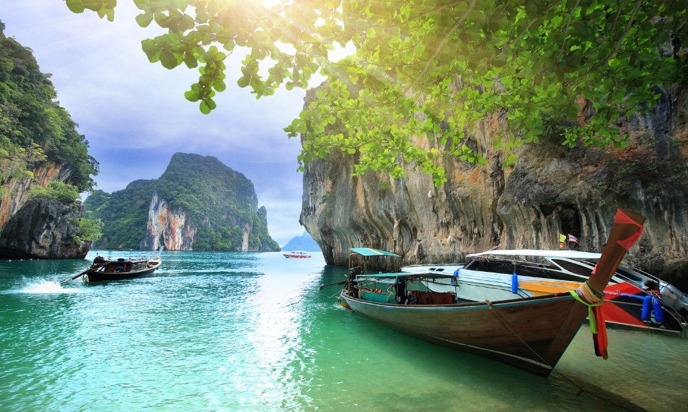 เรือหางยาวลอยอยู่บนน้ำสีฟ้าใสใกล้หน้าผาหินปูนและต้นไม้เขียวชอุ่มภายใต้แสงแดดจ้าในอ่าวเขตร้อน—เหมาะสำหรับการเที่ยวกระบี่และค้นพบสถานที่ท่องเที่ยวที่น่าทึ่งในกระบี่.