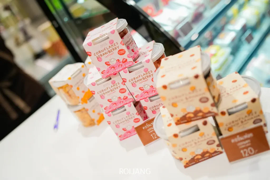 กล่องขนมคอร์นเฟลกส์ BEARHOUSE รสสตรอว์เบอร์รี่และช็อกโกแลตวางซ้อนกันอยู่บนเคาน์เตอร์สีขาว โดยมีตู้แช่เย็นแสดงสินค้าอยู่ด้านหลัง.