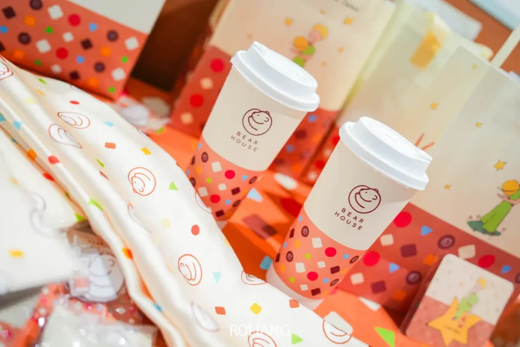 แก้วกาแฟแบบซื้อกลับบ้านสองใบที่มีโลโก้ BEARHOUSE ตั้งอยู่บนพื้นผิวสีส้มที่มีลวดลาย ล้อมรอบด้วยถุงตกแต่งและบรรจุภัณฑ์ที่มีสีสันสดใส.