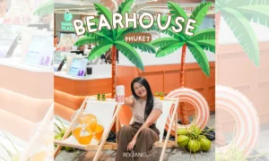 ผู้หญิงคนหนึ่งนั่งอยู่บนเก้าอี้ชายหาดถือเครื่องดื่มในมือ โพสท่าอยู่หน้าป้าย BEARHOUSE Phuket ที่สดใส รายล้อมไปด้วยต้นไม้ปาล์มและอุปกรณ์ชายหาดที่สนุกสนาน.