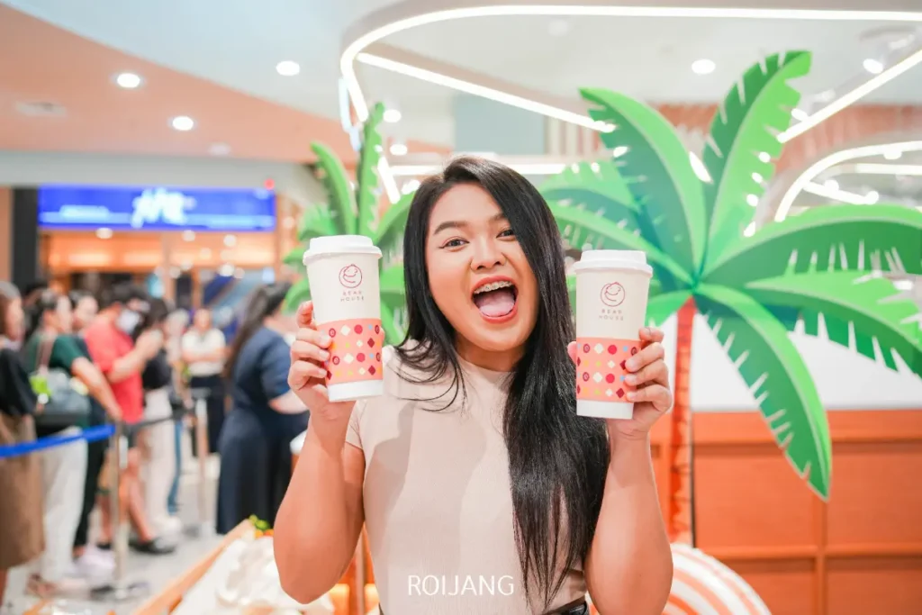 ผู้หญิงคนหนึ่งยิ้มขณะถือแก้วกาแฟสองแก้วในมือในบรรยากาศภายในที่สว่างไสว ตกแต่งด้วยต้นปาล์มเทียม และมีผู้คนอยู่ในฉากหลัง—อาจกำลังเพลิดเพลินกับของหวานจาก Simon Says Ice Cream.