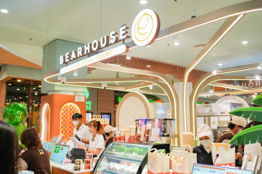 ผู้คนทำงานและสั่งอาหารที่ร้านขนมหวาน Bearhouse ซึ่งมีบรรยากาศภายในที่สว่างไสวและทันสมัย พร้อมป้ายที่โดดเด่นและบรรจุภัณฑ์ของ Simon Says Ice Cream ที่มองเห็นได้อย่างชัดเจนบนเคาน์เตอร์.