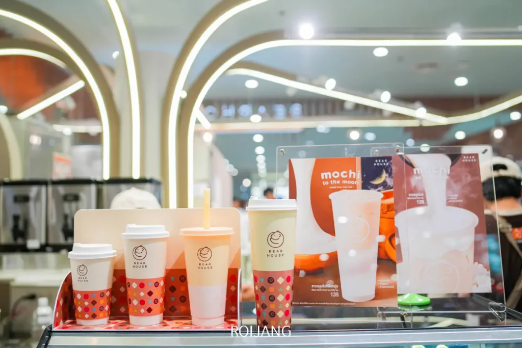 การจัดแสดงแก้วเครื่องดื่มแบรนด์ Bear House ขนาดต่างๆ และเมนูที่โปรโมตเครื่องดื่มโมจิ วางอยู่ข้างๆ ไอศกรีม Simon Says บนเคาน์เตอร์ภายในคาเฟ่ที่สว่างไสว.