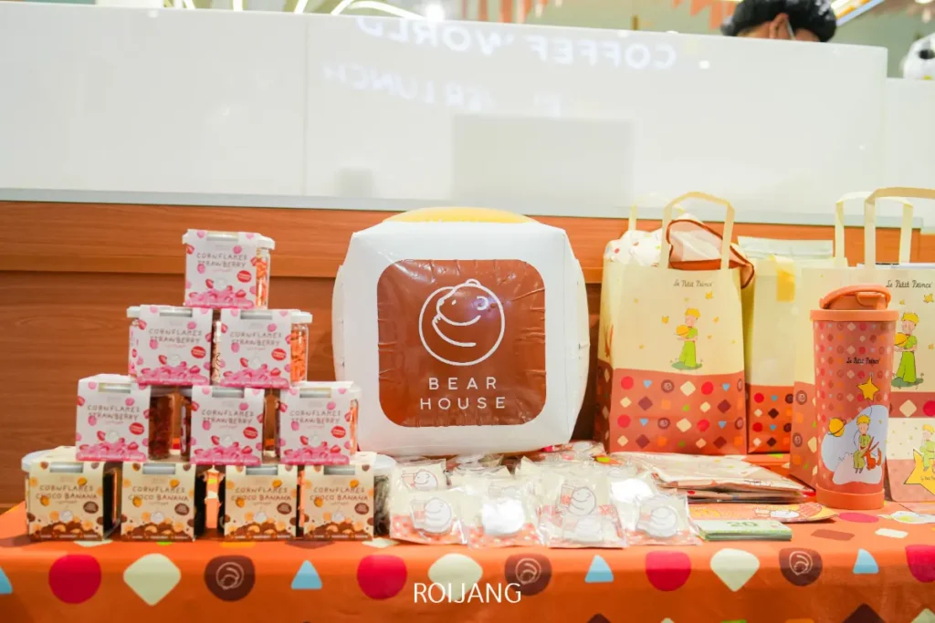 โต๊ะจัดแสดงสินค้า BEARHOUSE ประกอบด้วยกล่องซ้อนกัน หมอนใบใหญ่ กระเป๋าผ้า ถ้วยน้ำ และสินค้าบรรจุหีบห่อ จัดวางอย่างเป็นระเบียบบนผ้าสีส้ม.